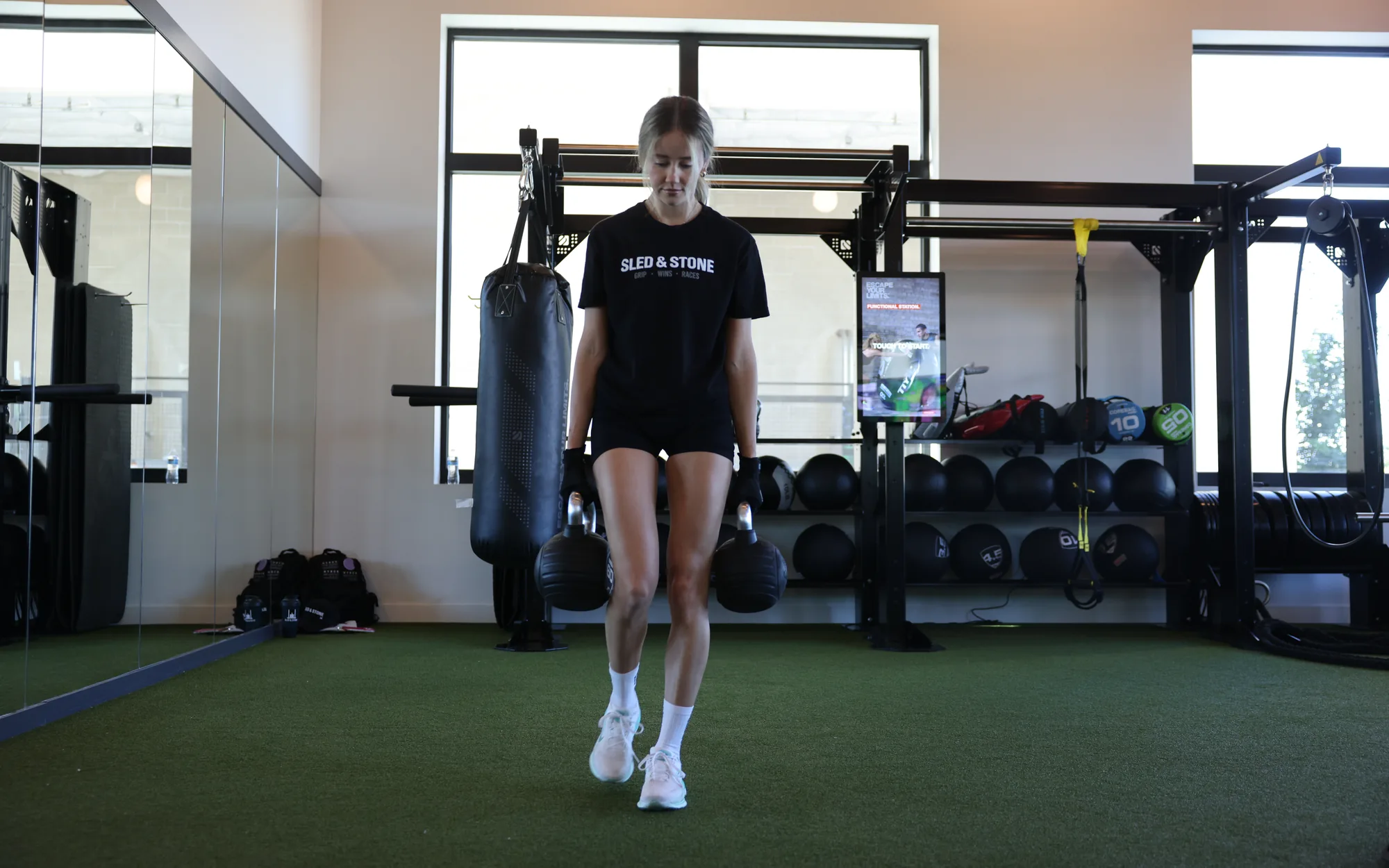 An athlete walking a farmers carry across a gym, a kettlebell in each gloved hand, in a Sled & Stone tee