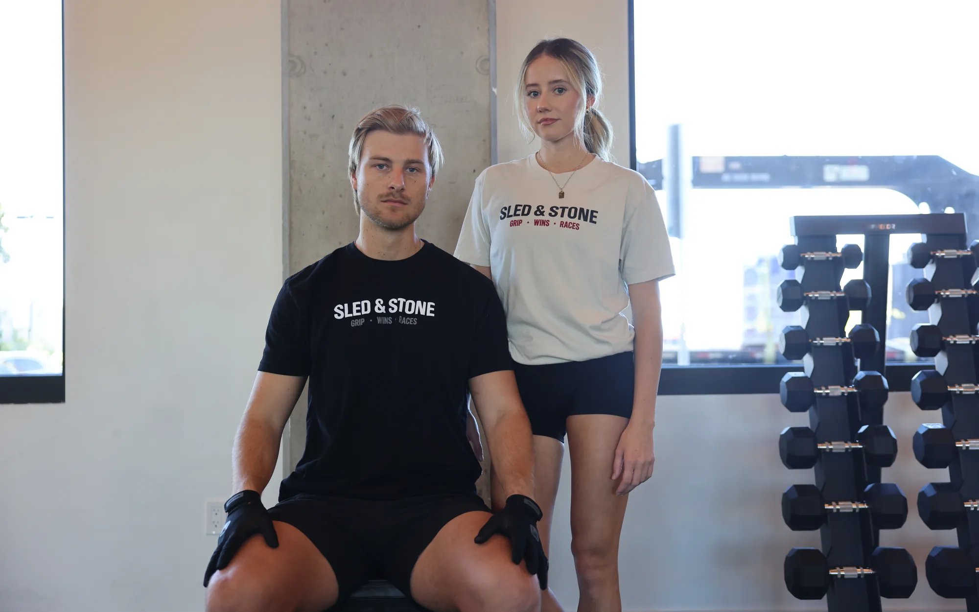 Two athletes in a gym in Sled & Stone tees and training shorts, one seated on a box wearing gloves