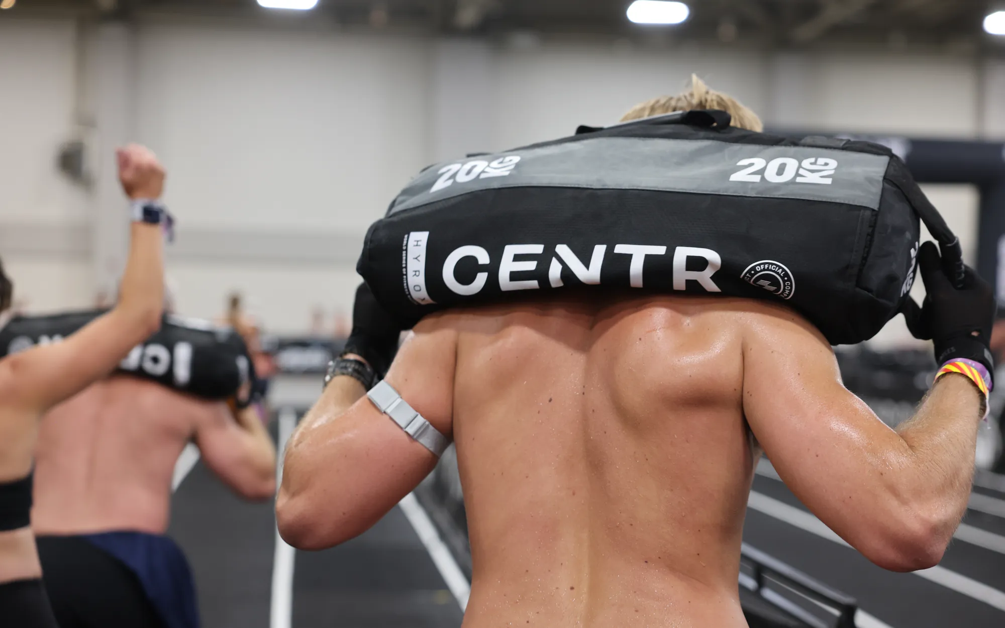 An athlete seen from behind with a 20kg HYROX sandbag across his shoulders, a gloved hand gripping each end, as other athletes lunge alongside