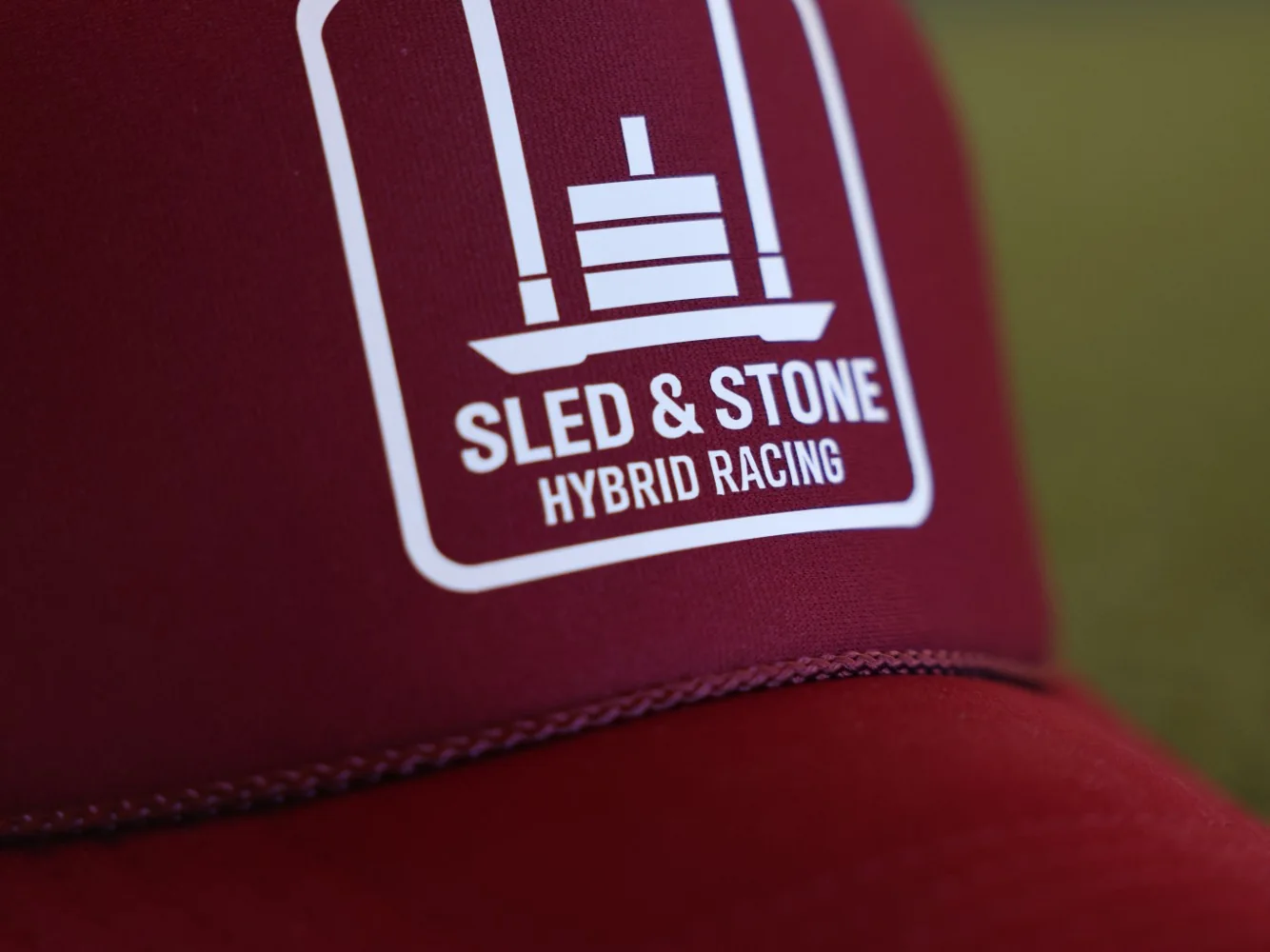 The red Hybrid Racing trucker, laid on turf, with the white Sled & Stone Hybrid Racing patch on the front — view 2