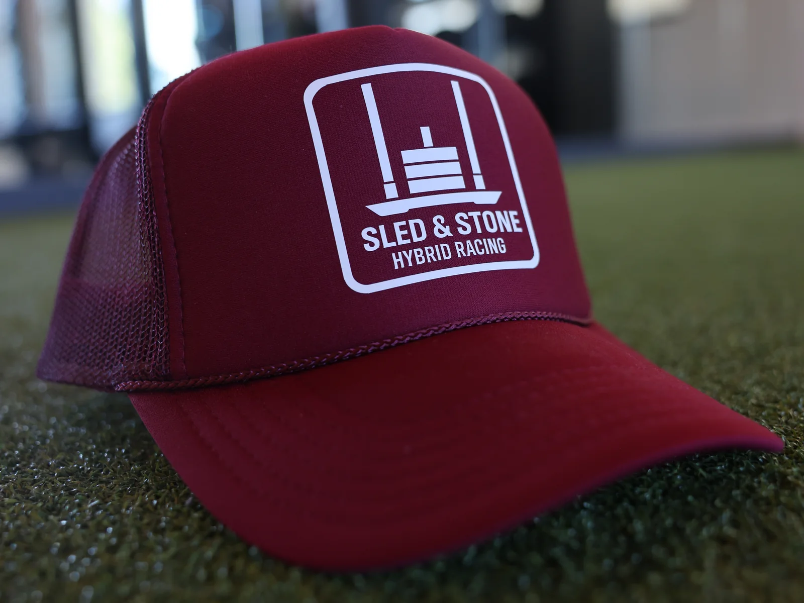 The red Hybrid Racing trucker, laid on turf, with the white Sled & Stone Hybrid Racing patch on the front