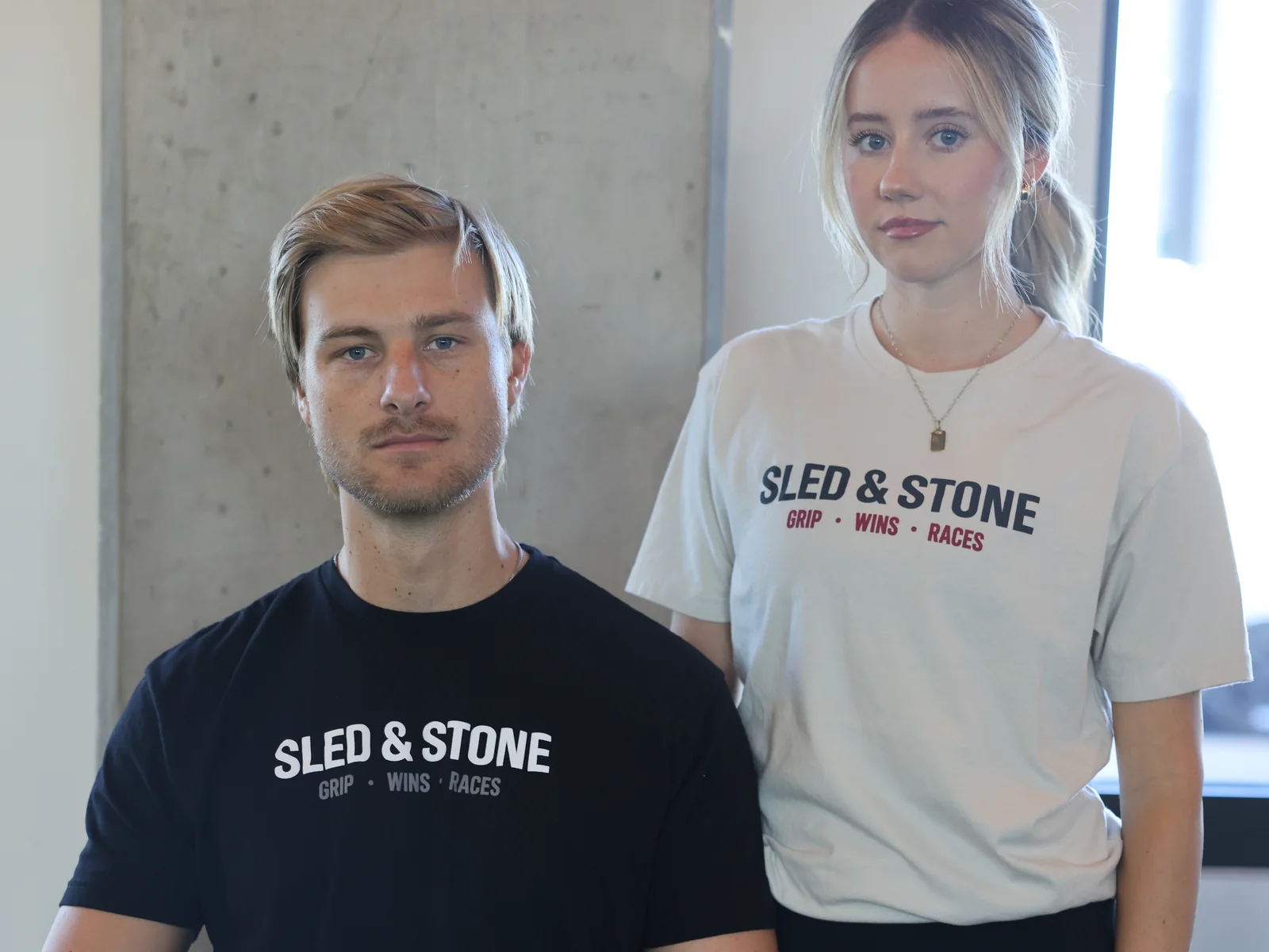 The Grip Wins Races tee in black and sand, worn side by side: Sled & Stone across the chest over Grip Wins Races